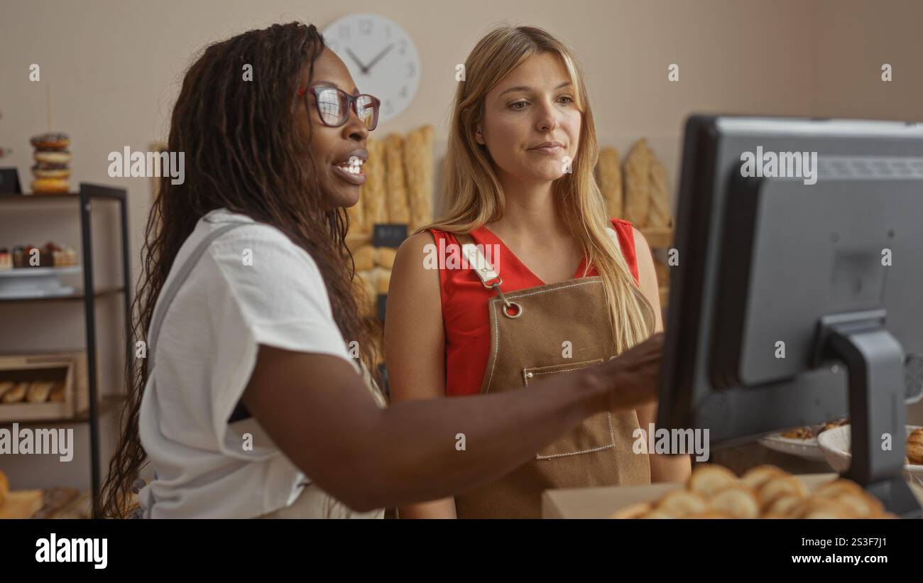 Zwei Frauen, Bäckerei-Arbeiter in Schürzen, stehen drinnen vor einem Computermonitor in einer Bäckerei, umgeben von Brot und Backwaren. Stockfoto