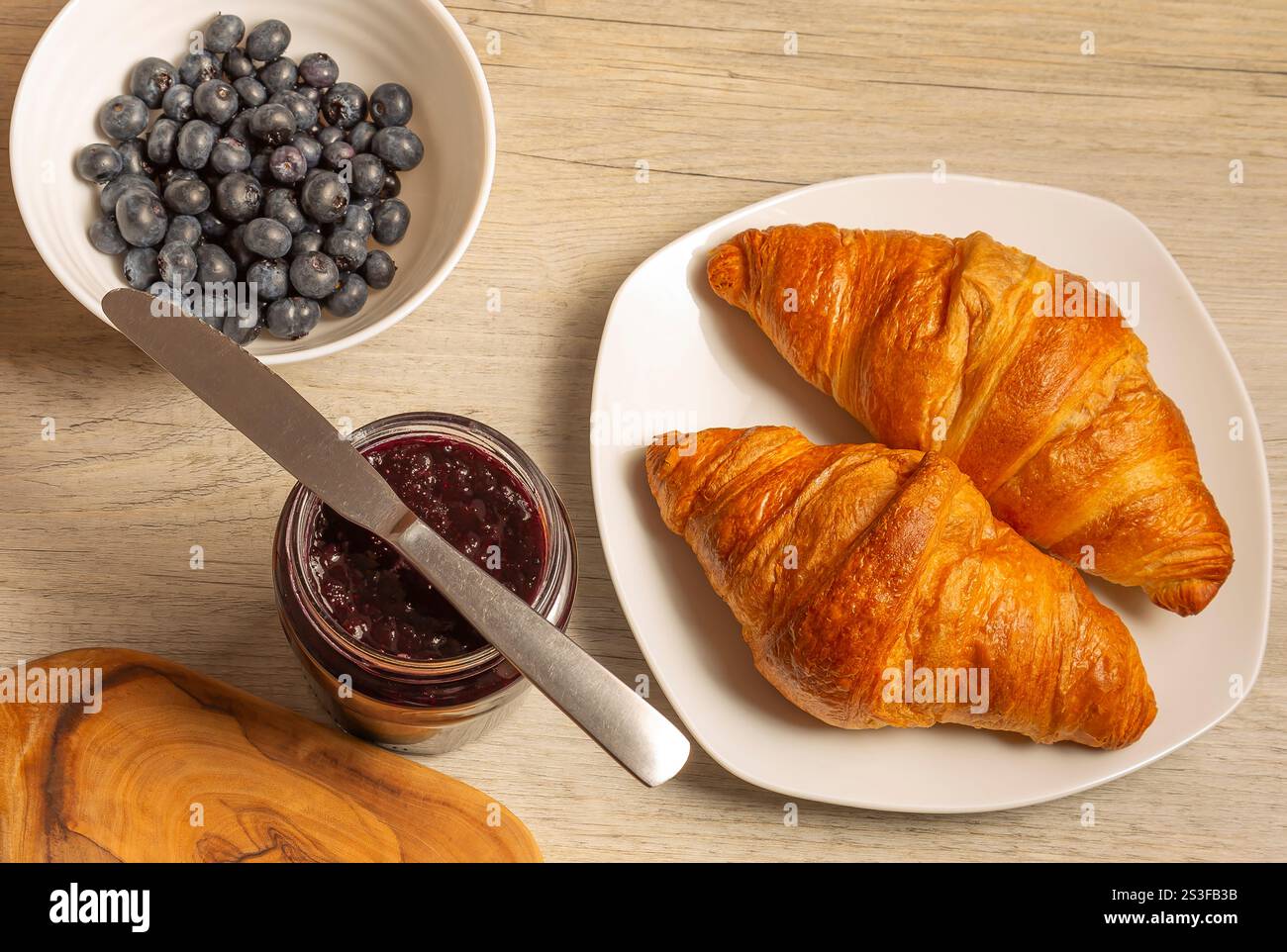 Zwei goldene Croissants auf einem weißen Teller, ein Glas mit dunklem Fruchtkonfitüre und eine Schüssel Heidelbeeren. Stockfoto