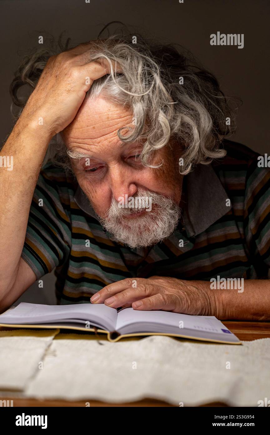 Ein älterer Mann mit grauen Haaren und gestreiftem Hemd sitzt an einem Tisch und konzentriert sich auf das Lesen eines Buches. Seine Hand liegt besinnlich auf dem Kopf. Stockfoto