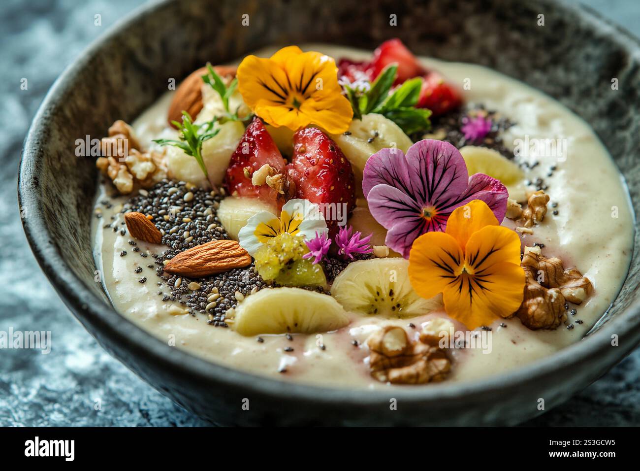 Eine lebendige Smoothie-Schüssel enthält geschnittene Bananen, Erdbeeren, Nüsse, Chiasamen und essbare Blumen, schön in einer Keramikschale angeordnet. Stockfoto