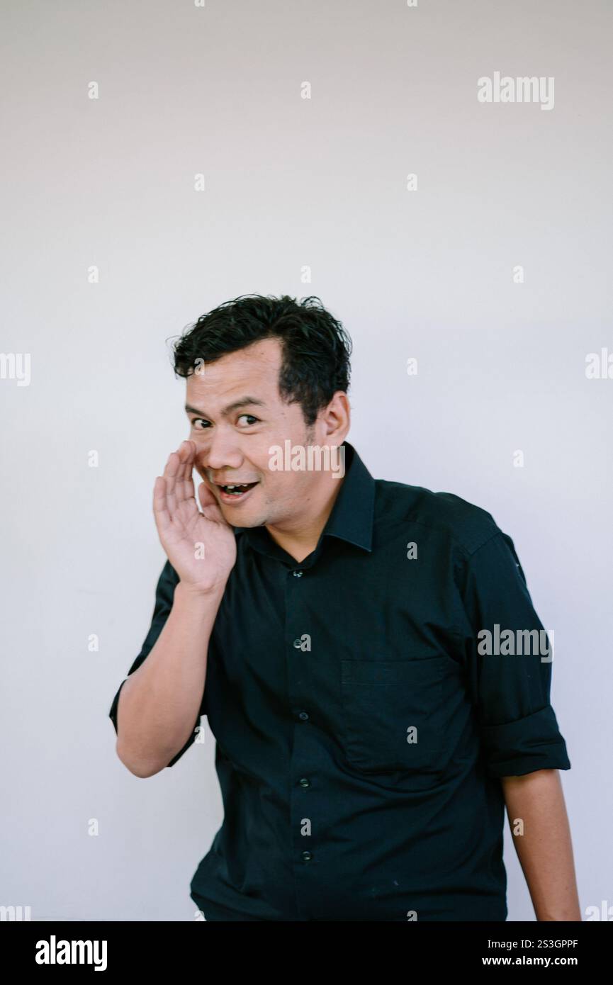Ein Mann in einem schwarzen Hemd, der eine verspielte Geste macht, flüstert mit der Hand in der Nähe seines Mundes vor einem weißen Hintergrund. Perfekt für das Konzept Stockfoto