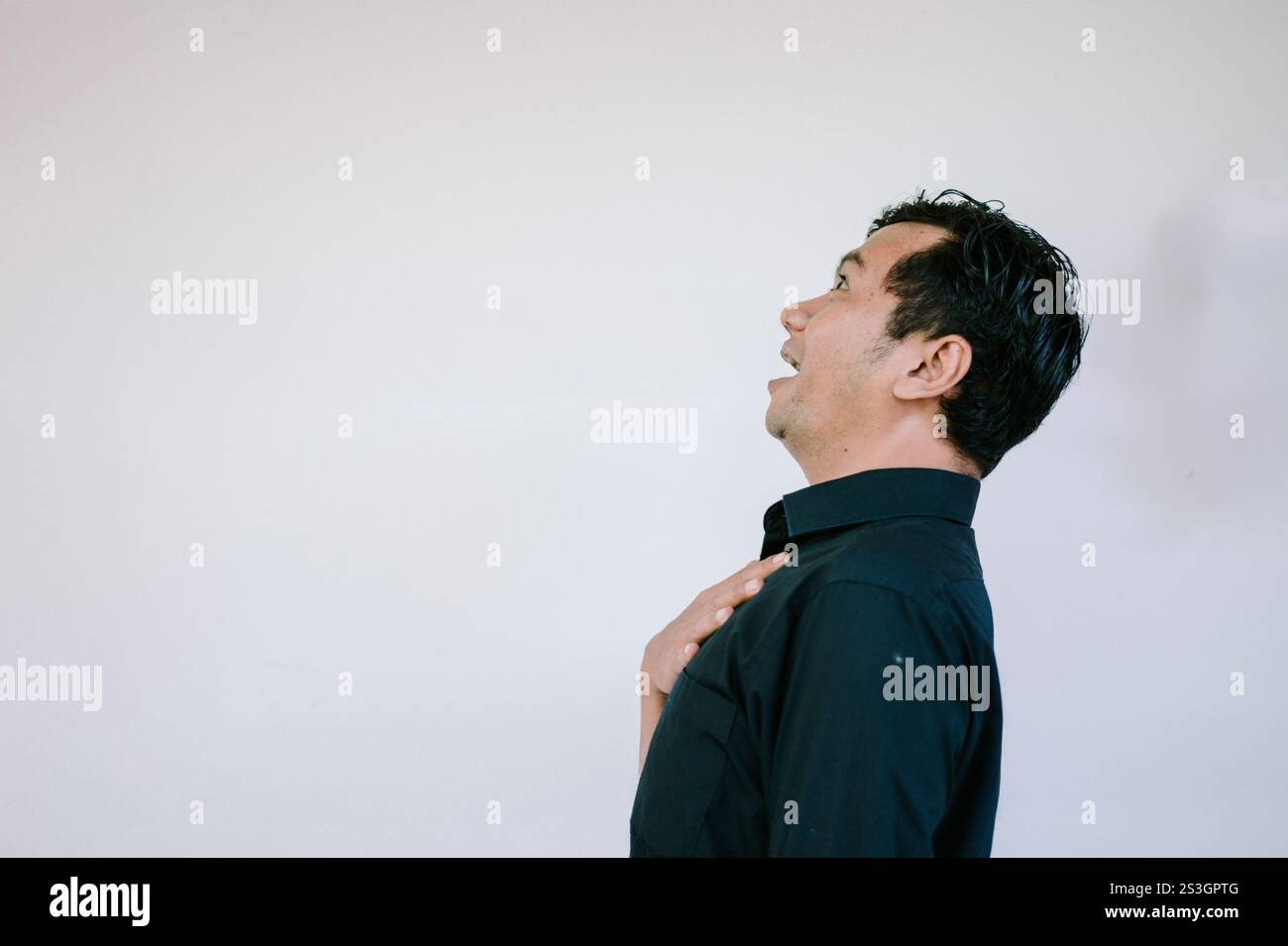 Ein Mann, der ein schwarzes Hemd trägt und mit einem überraschten oder verwunderten Ausdruck nach oben blickt, mit einer Hand auf der Brust, vor einem weißen Hintergrund. Ideal für Stockfoto