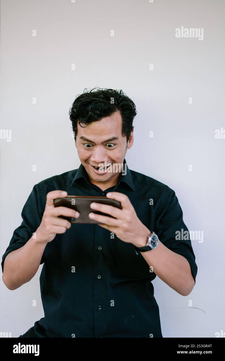 Ein Mann, der ein Smartphone mit einem aufgeregten und fokussierten Ausdruck hält und aufmerksam auf den Bildschirm starrt. Perfekt für Themen wie Gaming, Technologie oder Inte Stockfoto