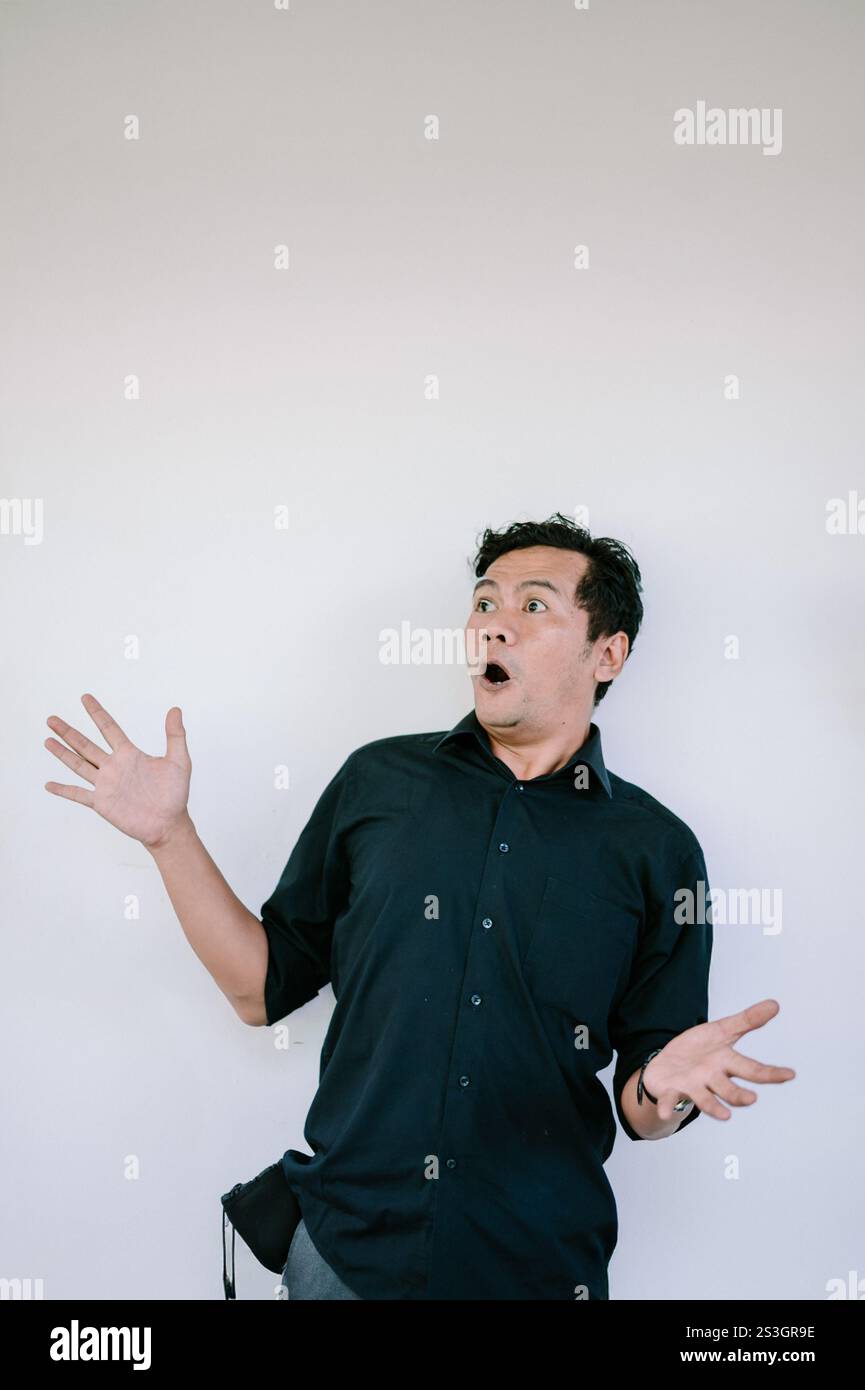 Ein Mann, der ein schwarzes Hemd trägt und mit einem überraschten oder verwunderten Ausdruck nach oben blickt, mit einer Hand auf der Brust, vor einem weißen Hintergrund. Ideal für Stockfoto