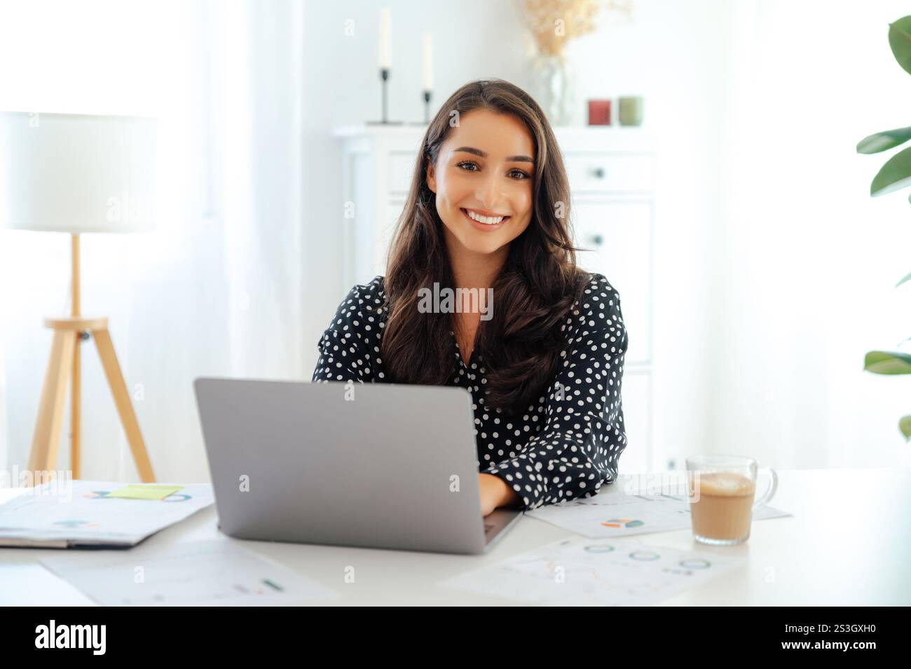 Glückliche, selbstbewusste, hübsche, brünette Frau, Büroangestellte, sitzt an einem weißen Schreibtisch mit einem Laptop, umgeben von Dokumenten und einer Tasse Kaffee im modernen Büro, blickt auf die Kamera und lächelt freundlich Stockfoto