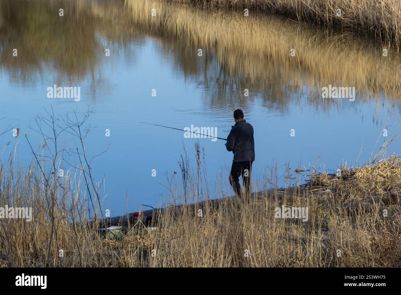 Ein Mann steht ruhig am Ufer des Wassers, am Ufer, umgeben von hohem Gras. Er konzentriert sich auf das Angeln im ruhigen Wasser Stockfoto