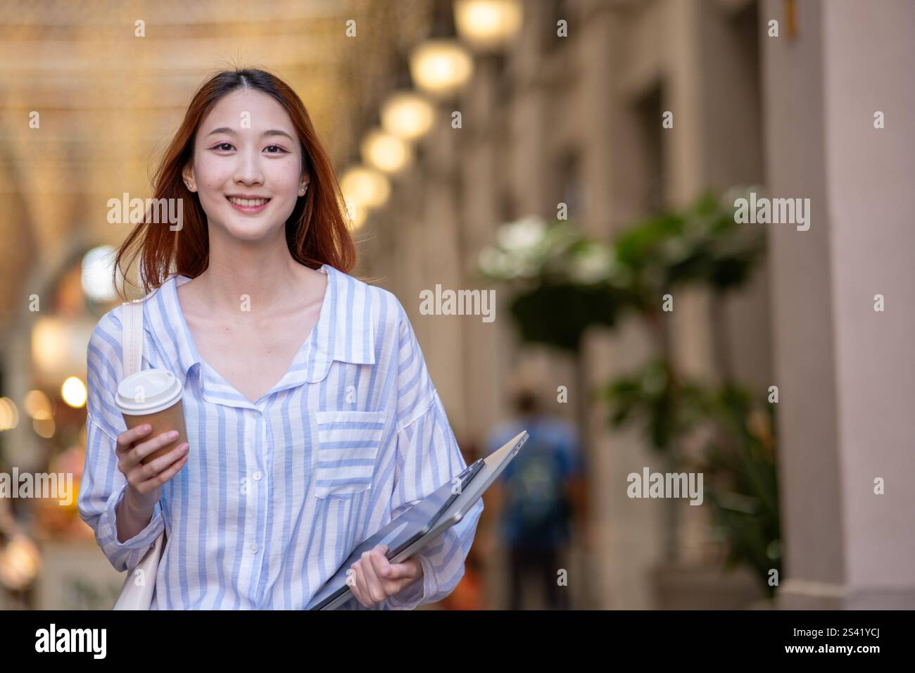 Eine Frau läuft die Straße hinunter und hält eine Tasse Kaffee und einen Stapel Papiere. Sie lächelt und ist gut gelaunt Stockfoto