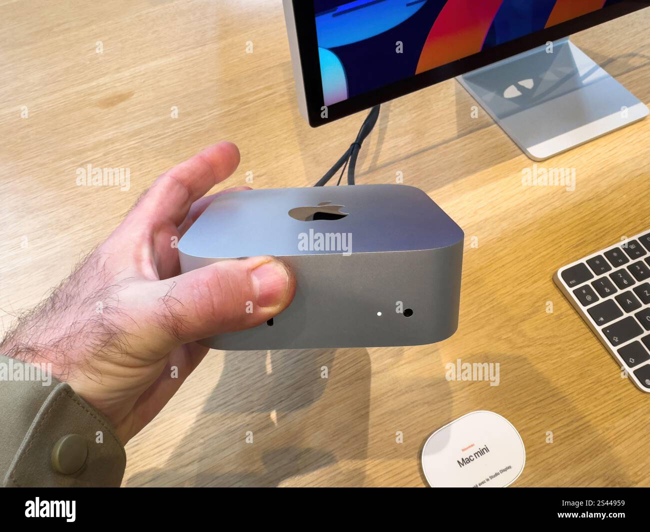 Paris, Frankreich - 10. Dezember 2024: Nahaufnahme einer Hand, die einen Apple Mac Mini in einem Geschäft hält, mit Monitor und Tastatur Stockfoto