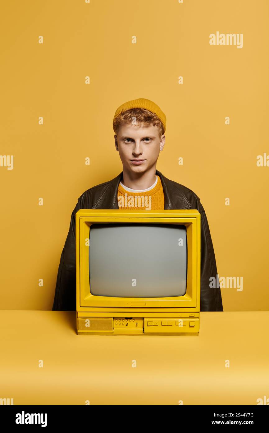 Ein modischer junger Mann mit rotem Haar zeigt seine Winterkleidung auf einem Retro-Fernseher in einem lebendigen Studio. Stockfoto