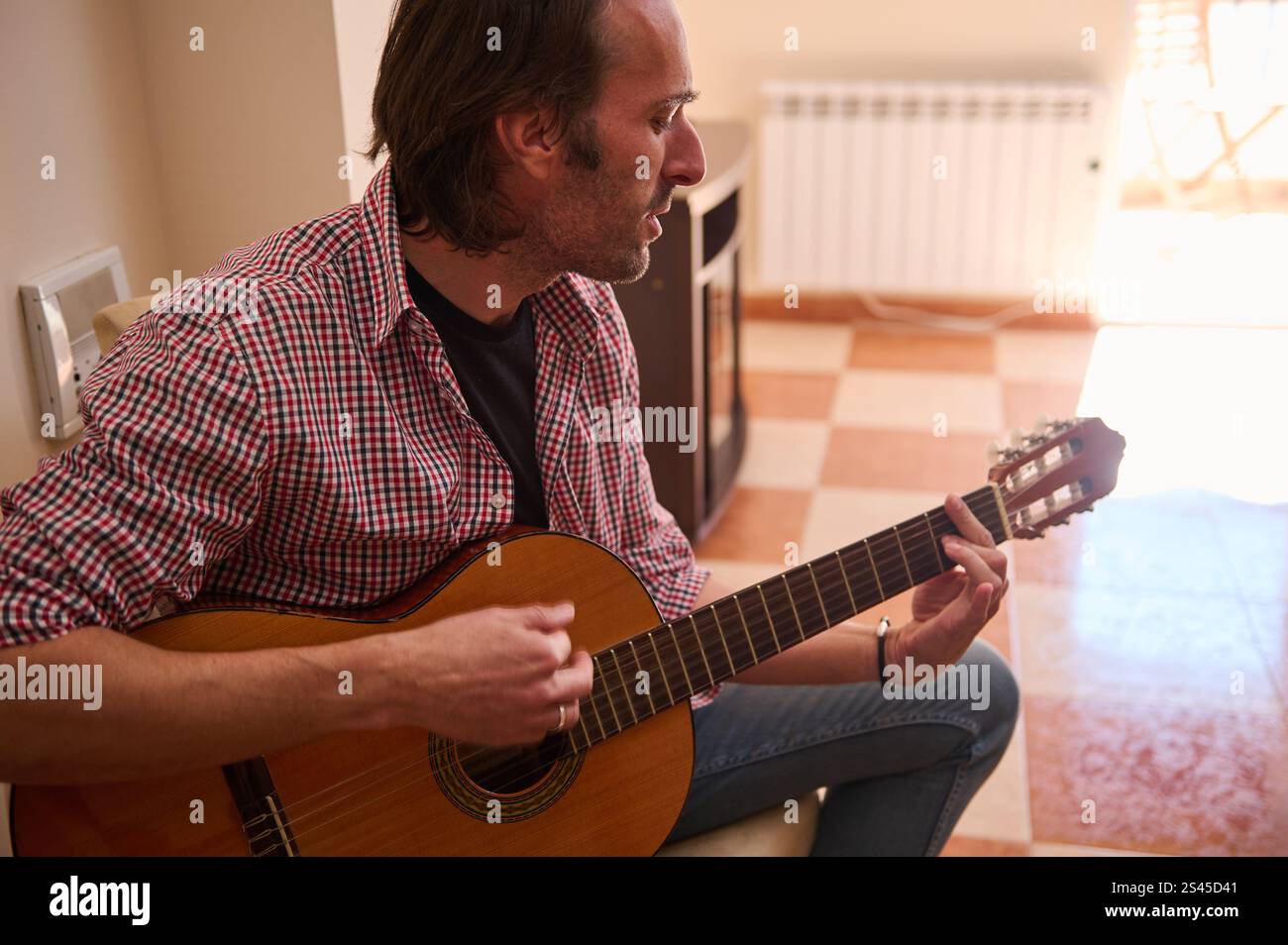Ein Mann sitzt und spielt Akustikgitarre in einem hellen, gemütlichen Raum mit eindringendem Sonnenlicht. Die Szene zeigt Kreativität und Entspannung, perfekt für Stockfoto