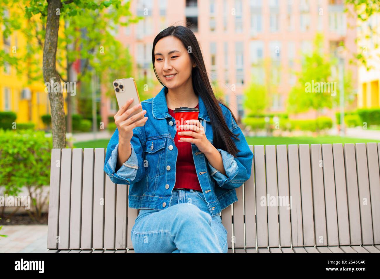 Asiatische Koreanerin, die ein Smartphone benutzt und auf einer Bank im öffentlichen Stadtgarten sitzt, eine Einweg-Kaffeetasse hält Stockfoto