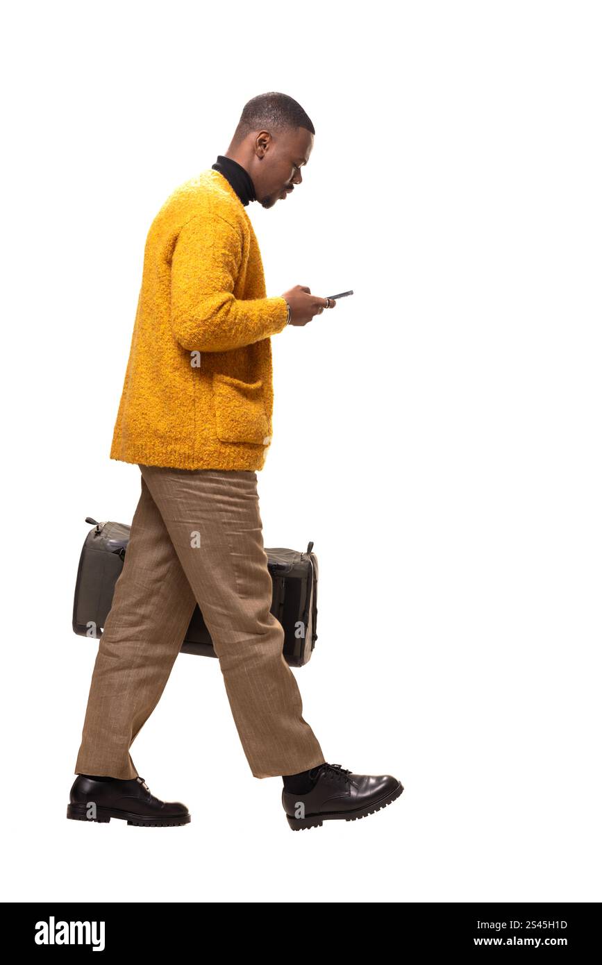 Ein junger Mann in einem stilvollen gelben Pullover und einer braunen Hose geht in einem Studio, hält ein Smartphone und eine Tasche und symbolisiert moderne Geschäftsmobilität und ef Stockfoto