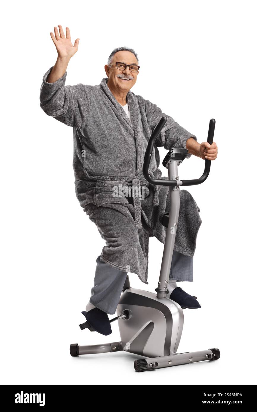 Reifer Mann in einem Bademantel, der auf einem Fitnessrad fährt und isoliert auf weißem Hintergrund winkt Stockfoto