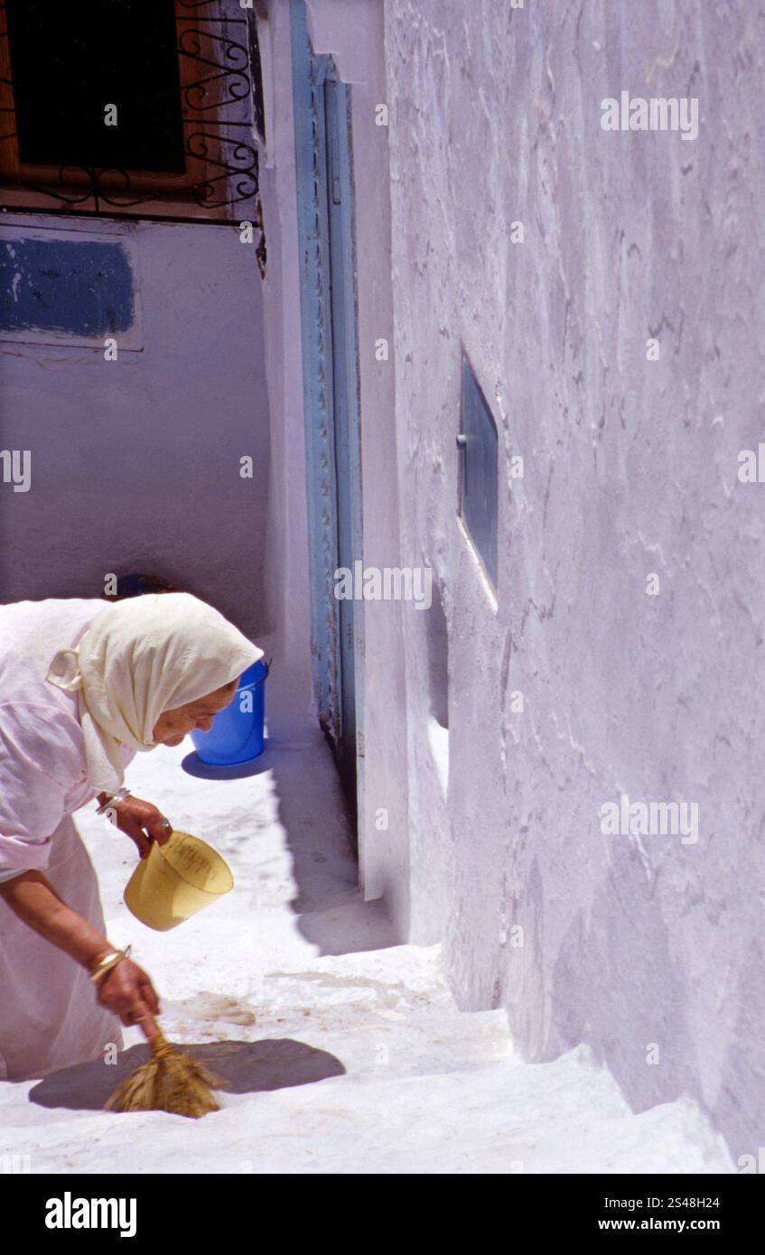 Frau, die eine weiß getünchte Straße im andalusischen Viertel Chefchaouen, Marokko, durchstreift, das für seine blau gestrichenen Gebäude und seinen traditionellen Charme bekannt ist. Stockfoto