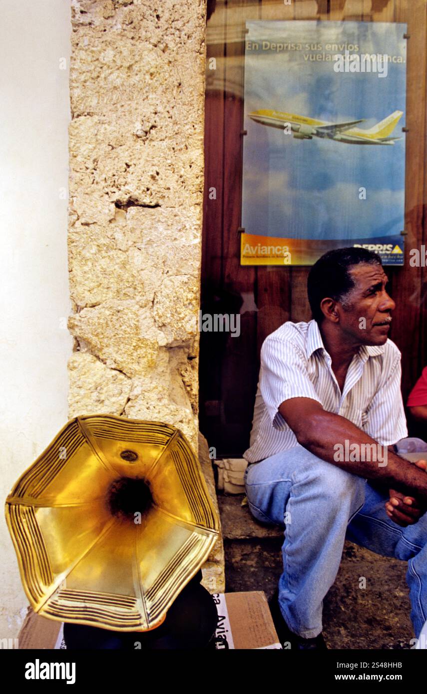 Grammophon-Verkäufer auf einem Flohmarkt in Cartagena, Kolumbien, sitzt neben einem goldenen Horn-Grammophon mit einer Avianca-Anzeige im Hintergrund. Stockfoto