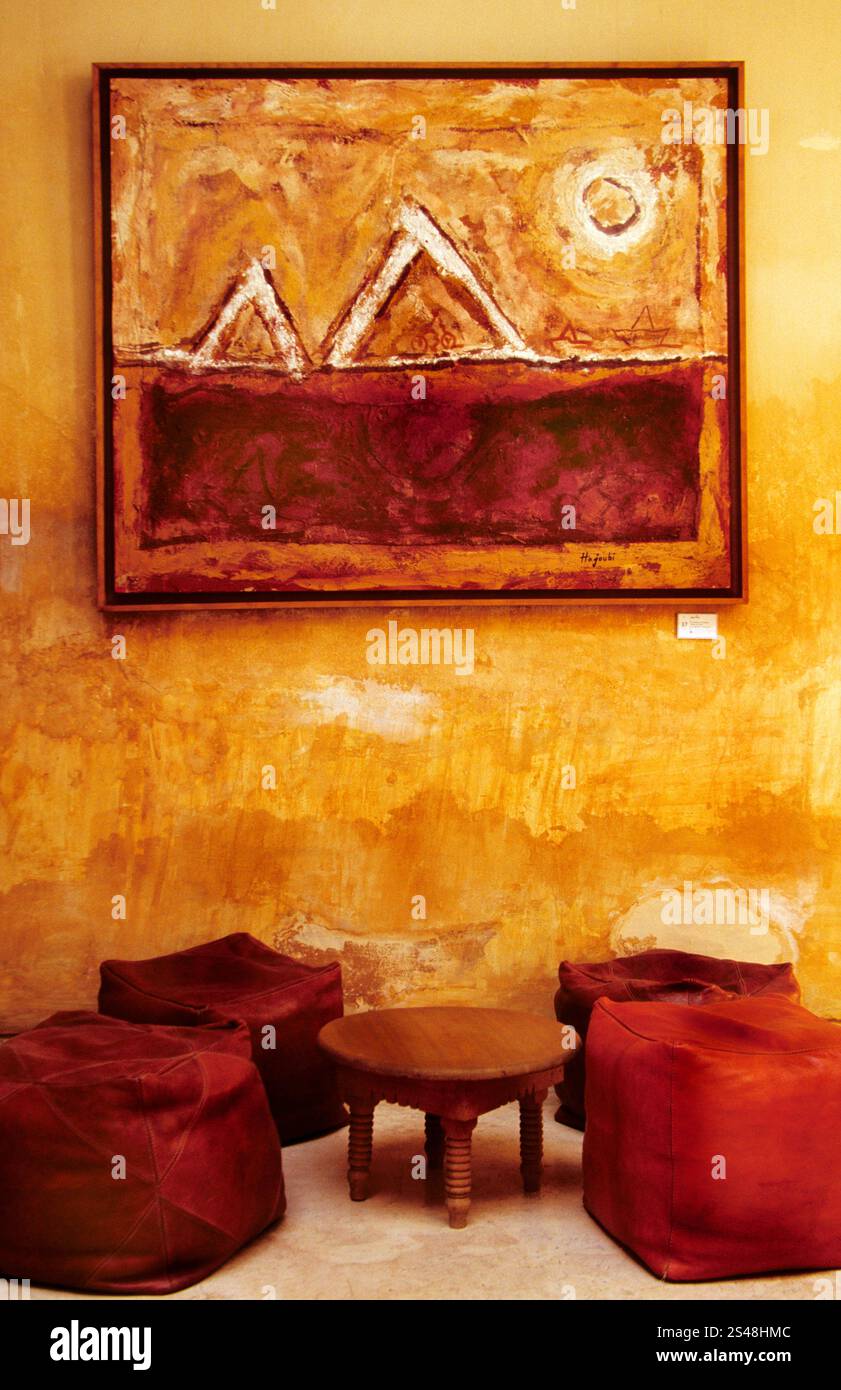 Abstrakte Malerei mit geometrischen Formen in einem Riad in Marrakesch, Marokko, mit einem rustikalen Sitzbereich mit Poufs und einem Holztisch. Stockfoto
