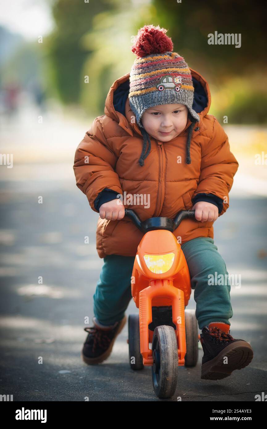 Ein kleines Kind fährt mit einem orangen Dreirad auf einem Parkweg, gekleidet in einer braunen Jacke und Strickmütze, und lächelt fröhlich, während die Sonne hell scheint Stockfoto