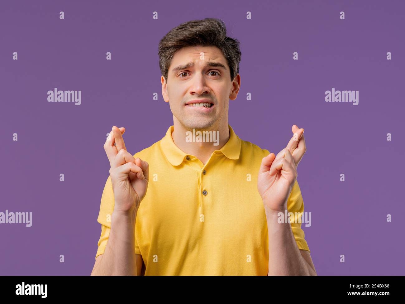 Junger Mann, der mit gekreuzten Fingern auf violettem Hintergrund betet. Der Typ fleht jemanden an, befriedigt Wünsche, hilft mit, betet um Glück in der Prüfung. Hohe Qualität Stockfoto