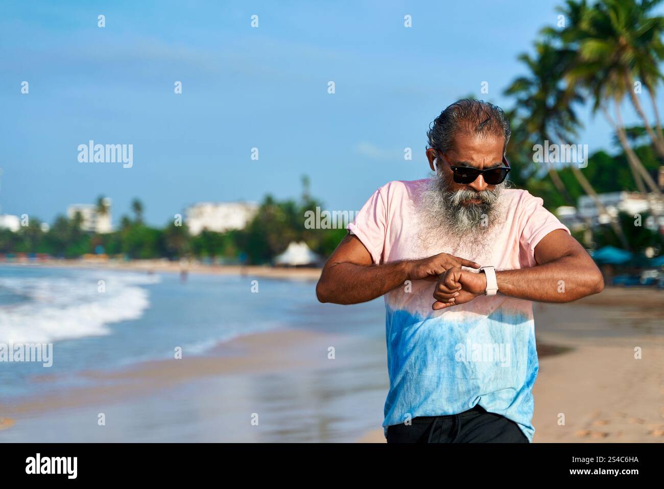Senior-Läufer aus Südasien am Strand mit Smartwatch und schnurlosen Ohrstöpseln. Ein bärtiger Mann überprüft den Fitness-Tracker während des morgendlichen Joggs. Moderne Technik für die Gesundheit Stockfoto