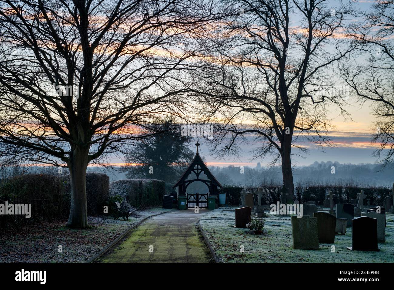 Grabsteine, Lychentore und Bäume am Abend Frost auf einem Friedhof in der Abenddämmerung. Kings Sutton, Northamptonshire, England. Stockfoto