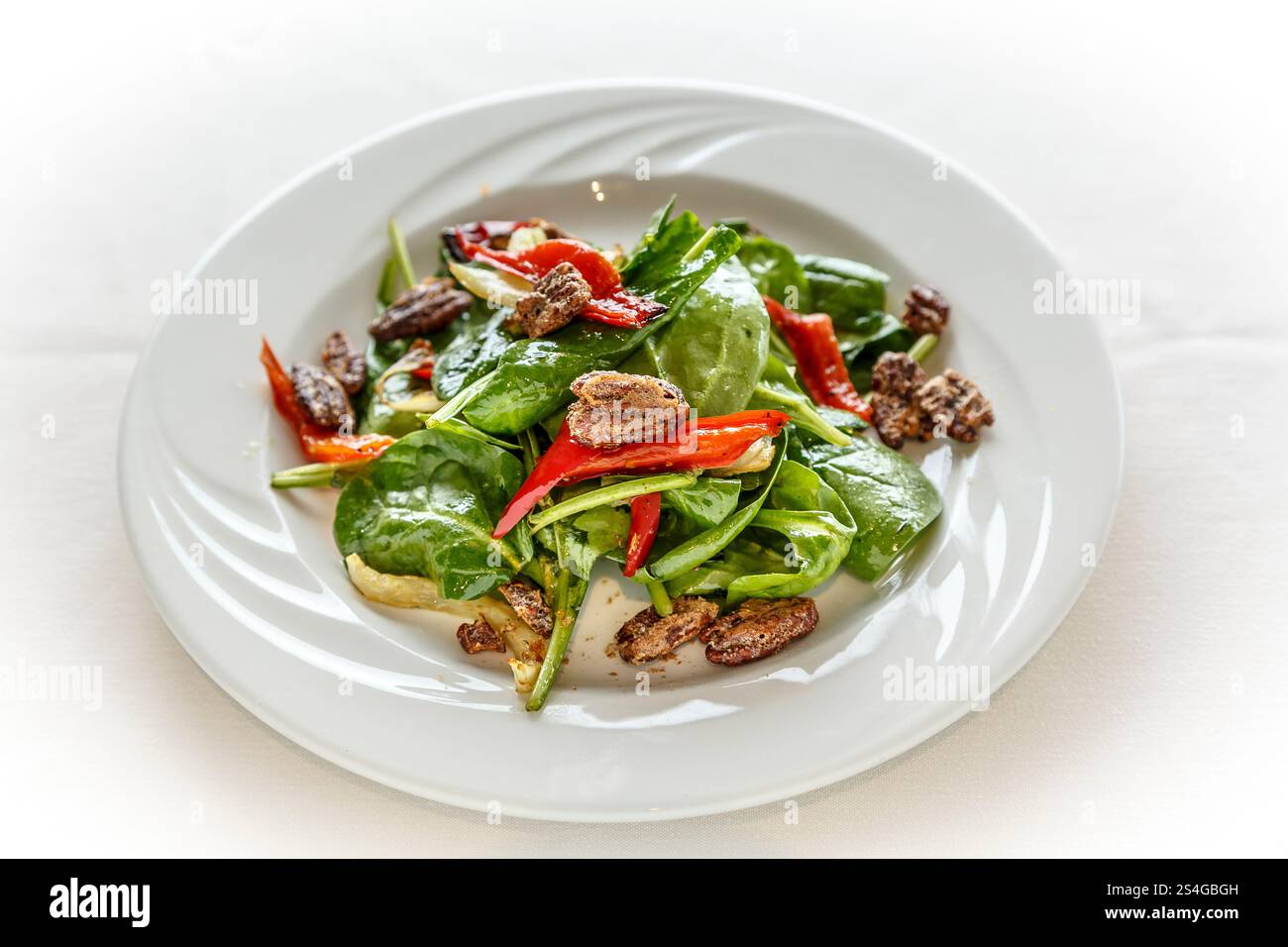 Ein Salat mit Spinat, Paprika und Nüssen auf einem weißen Teller. Der Salat wird auf einer weißen Tischdecke serviert Stockfoto