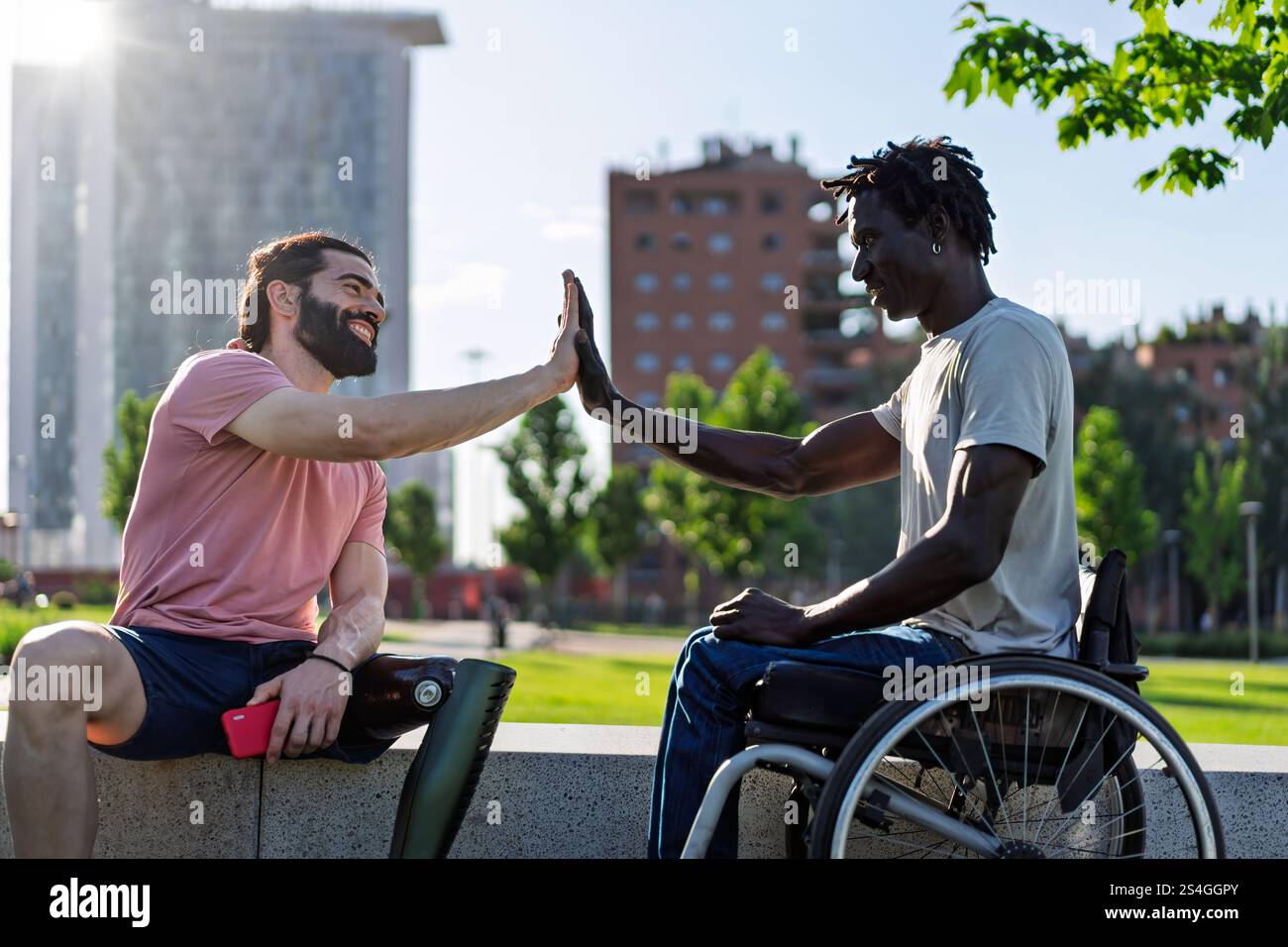 Zwei Freunde, einer mit einer Beinprothese und der andere im Rollstuhl, High-Fiving in einem Stadtpark. Das Bild vermittelt Empowerment, Diversität und Pommes Frites Stockfoto