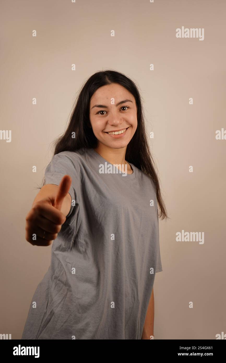 Fröhliches Mädchen hält eine Hand mit Daumen nach oben auf neutralem hellem Hintergrund. Ihre entspannte Pose bringt Zustimmung zum Ausdruck. Positiver Moment, der mit einer Hand erfasst wird Stockfoto