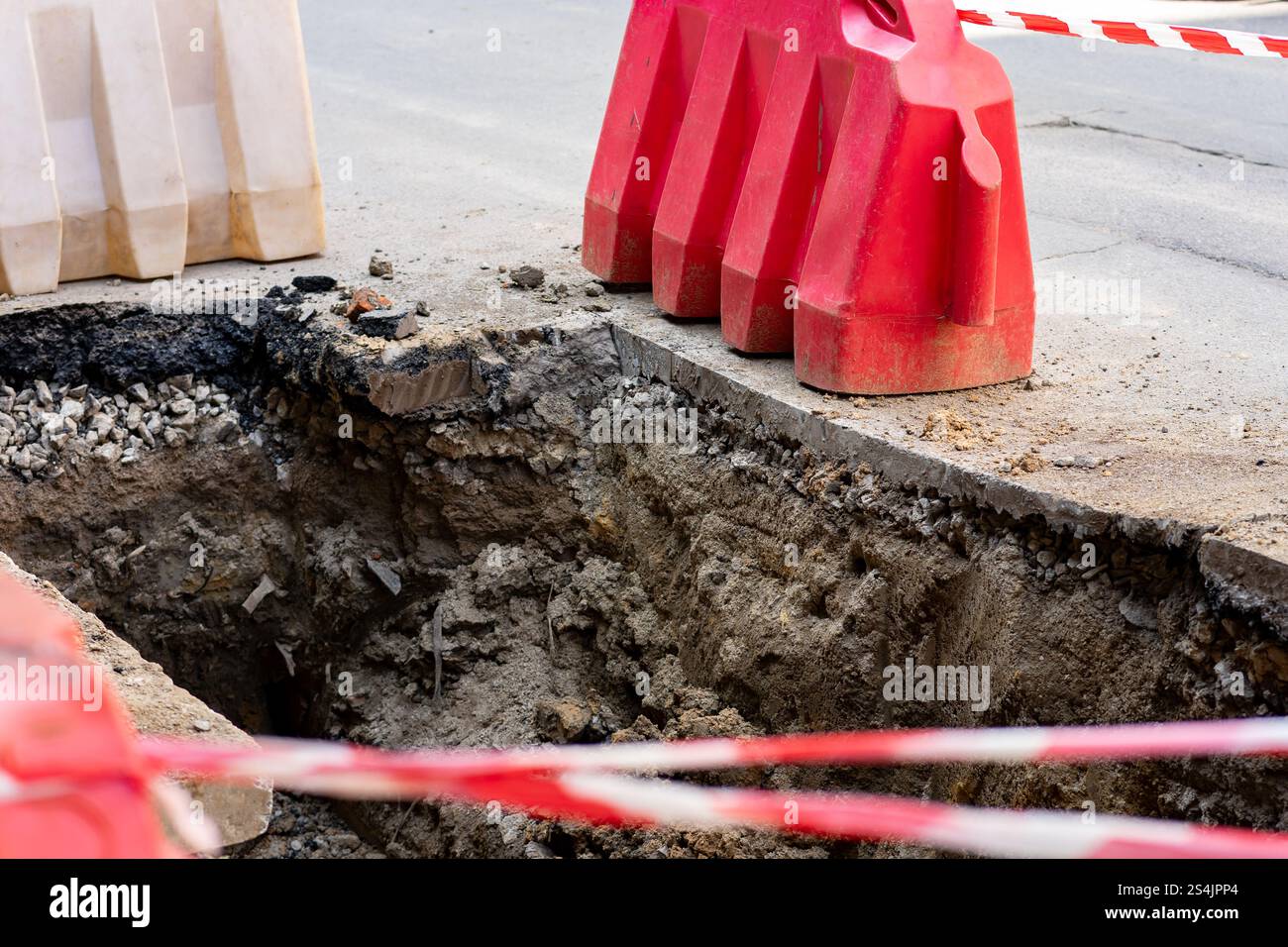 Die Arbeiter heben einen tiefen Graben auf der Straße aus, der von Sicherheitsbarrieren umgeben ist. Rotes und weißes Band signalisiert einen Gefahrenbereich und sorgt so für Sicherheit beim Pass Stockfoto