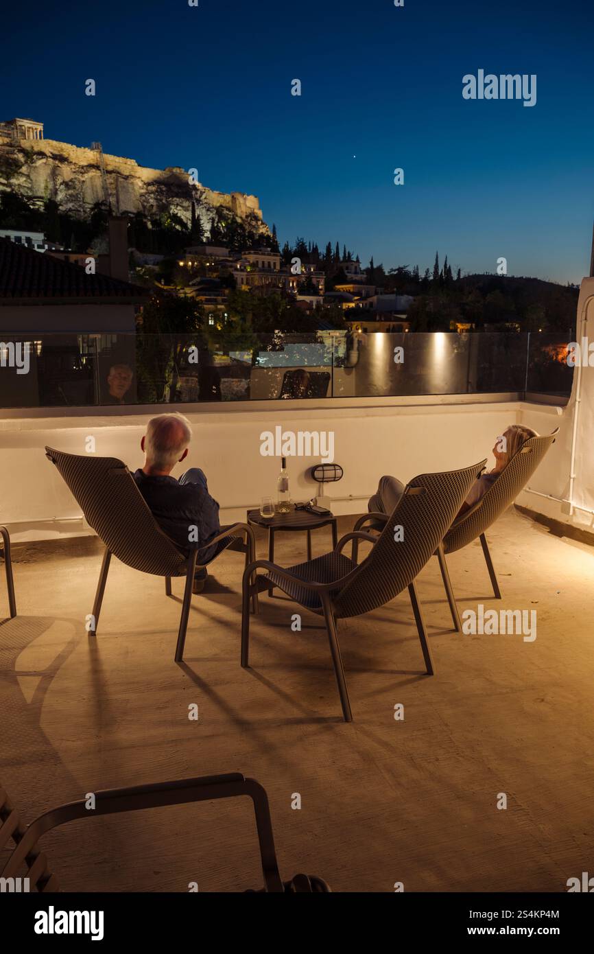 Ein Mann und eine Frau sitzen mit einer Flasche Wein in der Abenddämmerung in Liegestühlen auf einer Dachterasse in Athen und schauen auf die beleuchtet Stockfoto