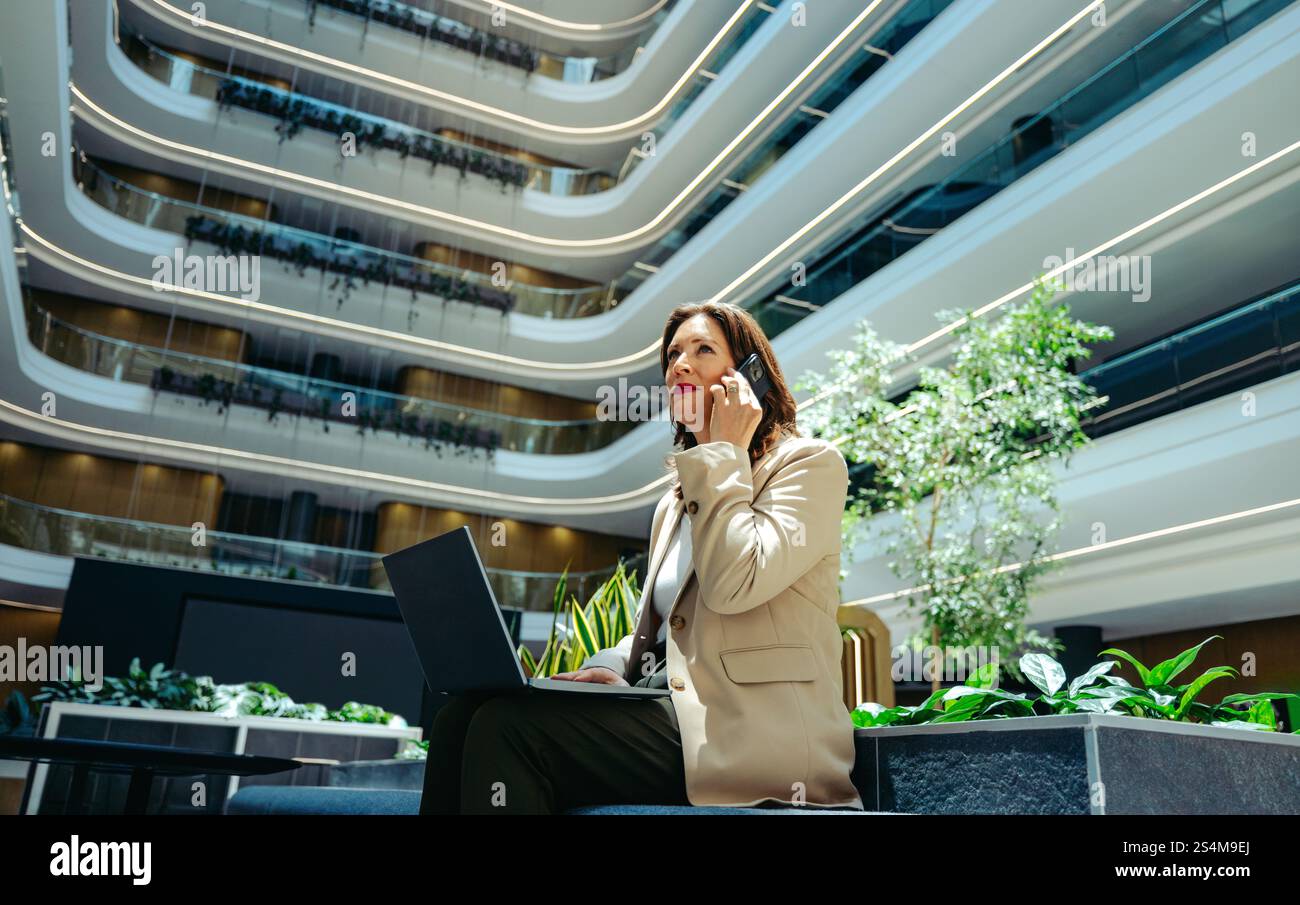 Eine Geschäftsfrau führt einen Anruf, während sie in einem modernen Innenhof sitzt. Die umliegende Architektur ist elegant und modern, mit lu Stockfoto