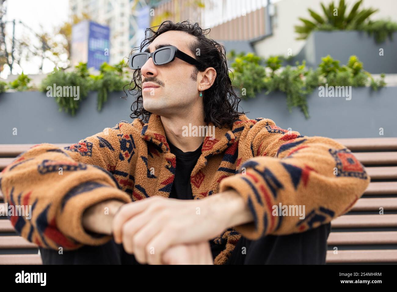 Ein modischer Mann mit Sonnenbrille und gemusterter Jacke sitzt draußen auf einer Bank und strahlt eine entspannte und selbstbewusste Atmosphäre in einer urbanen Umgebung aus Stockfoto