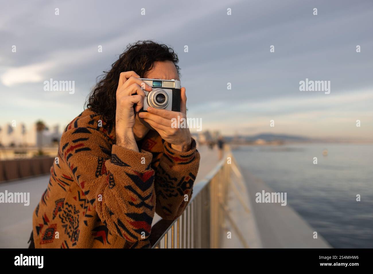 Lässiger Moment eines jungen Mannes, der die Fotografie am malerischen Ufer genießt. Er trägt einen gemütlichen, gemusterten Pullover und hält eine Kamera vor einer ruhigen Kulisse Stockfoto