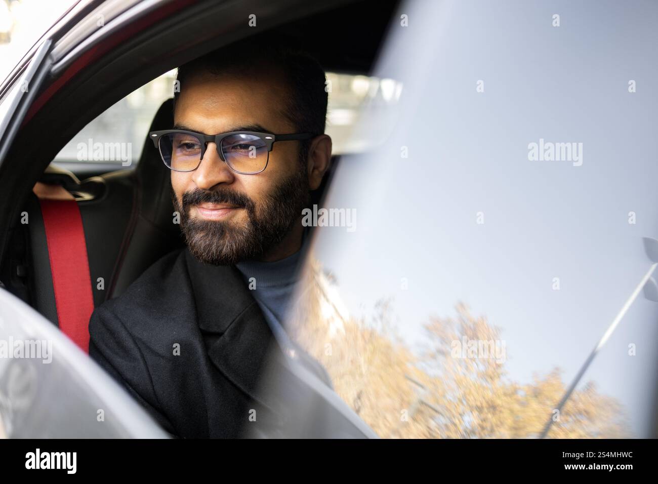 Ein Indianer, der in einem dunklen Wintermantel und einer Brille gekleidet ist, wird während der Fahrt mit seinem Auto sanft lächelnd gesehen, was ein Gefühl von Zufriedenheit und Komfort vermittelt. Stockfoto