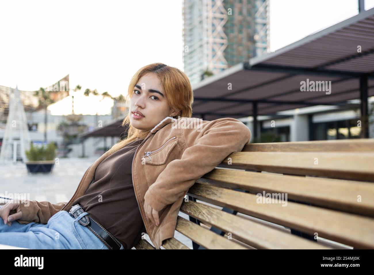 Gemischte Rassenfrau mit hellen Haaren, brauner Jacke, lässig auf einer Holzbank in einer modernen städtischen Umgebung mit Gebäuden und grüner Stockfoto