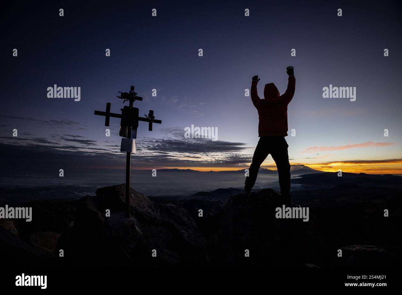 Ein nicht erkennbarer Mann steht mit erhobenen Armen nahe einem Kreuz mit Silhouette bei Sonnenaufgang auf einem zerklüfteten Berggipfel, was einen Moment des Triumphes und des Gipfels hervorhebt Stockfoto