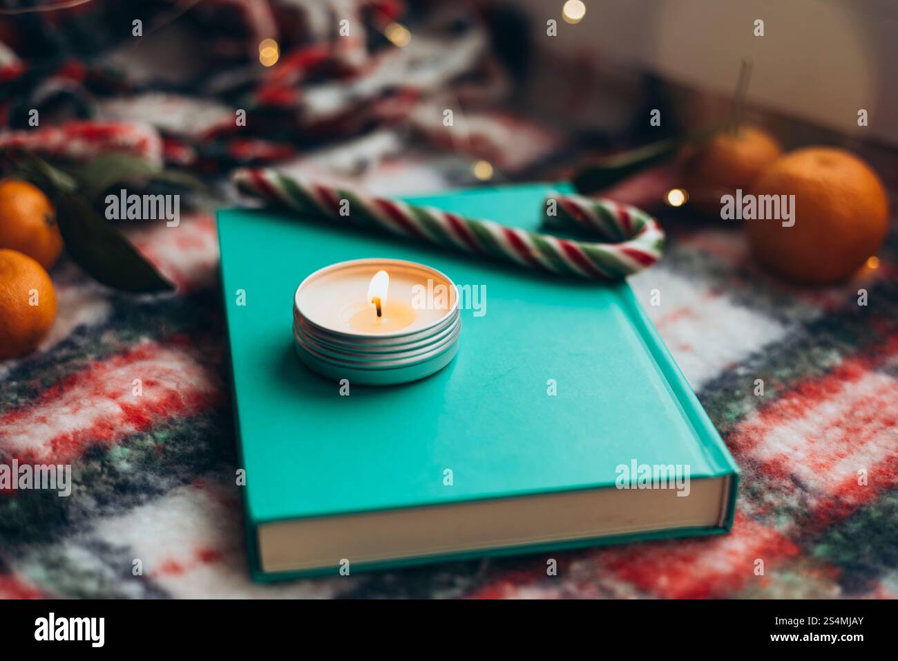 Friedliche Weihnachtskulisse mit einem Petrol-Buch und beleuchteter Kerze auf einer farbenfrohen festlichen Decke, umgeben von einem Zuckerrohr und Mandarinen-Orangen. Stockfoto