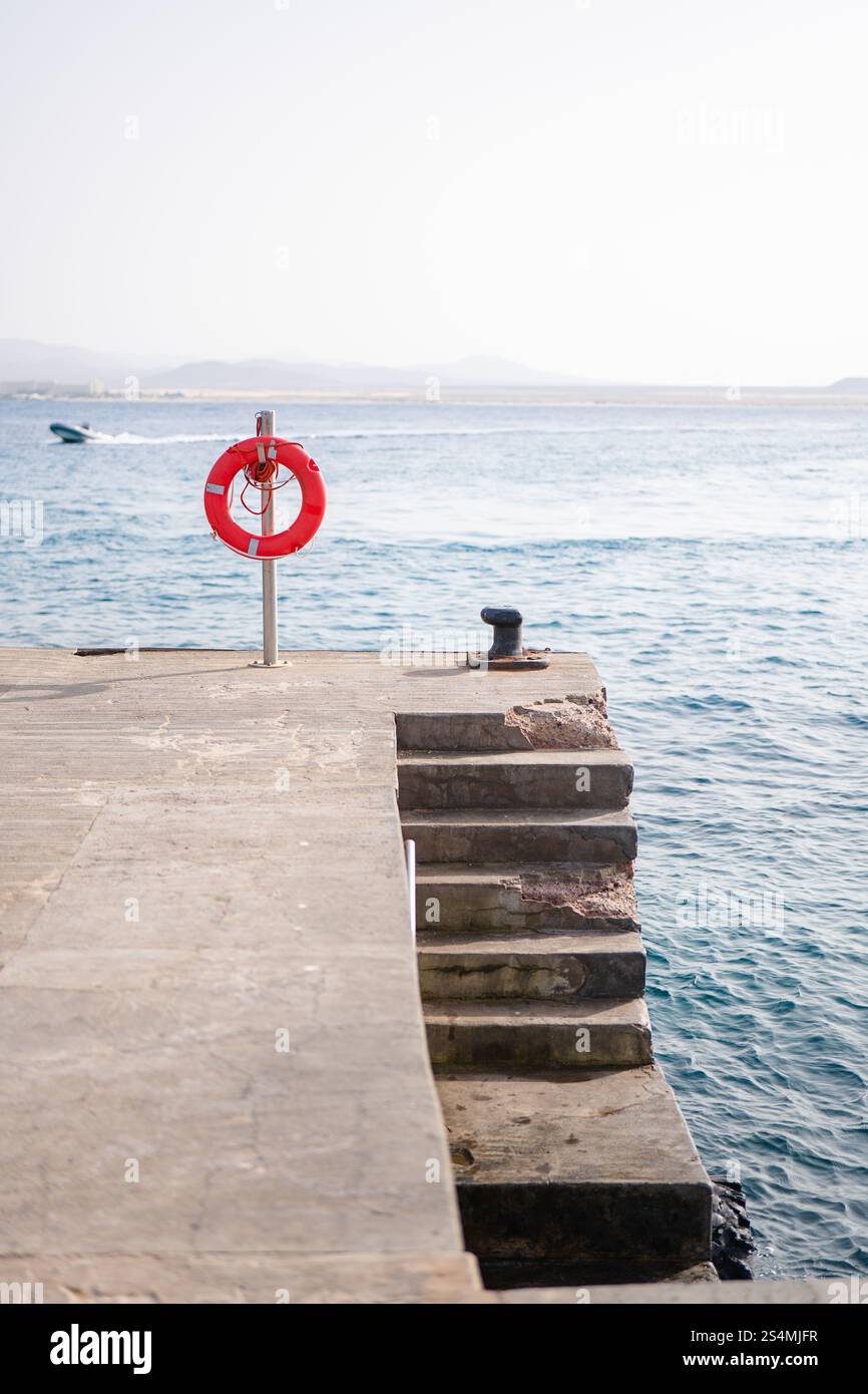 Eine ruhige Hafenszene mit einem leuchtend roten Rettungsschirm auf einem verwitterten Dock, mit Blick auf das ruhige Meer, Ein weit entferntes Boot erzeugt sanfte Wellen unter einem klaren Wasser Stockfoto