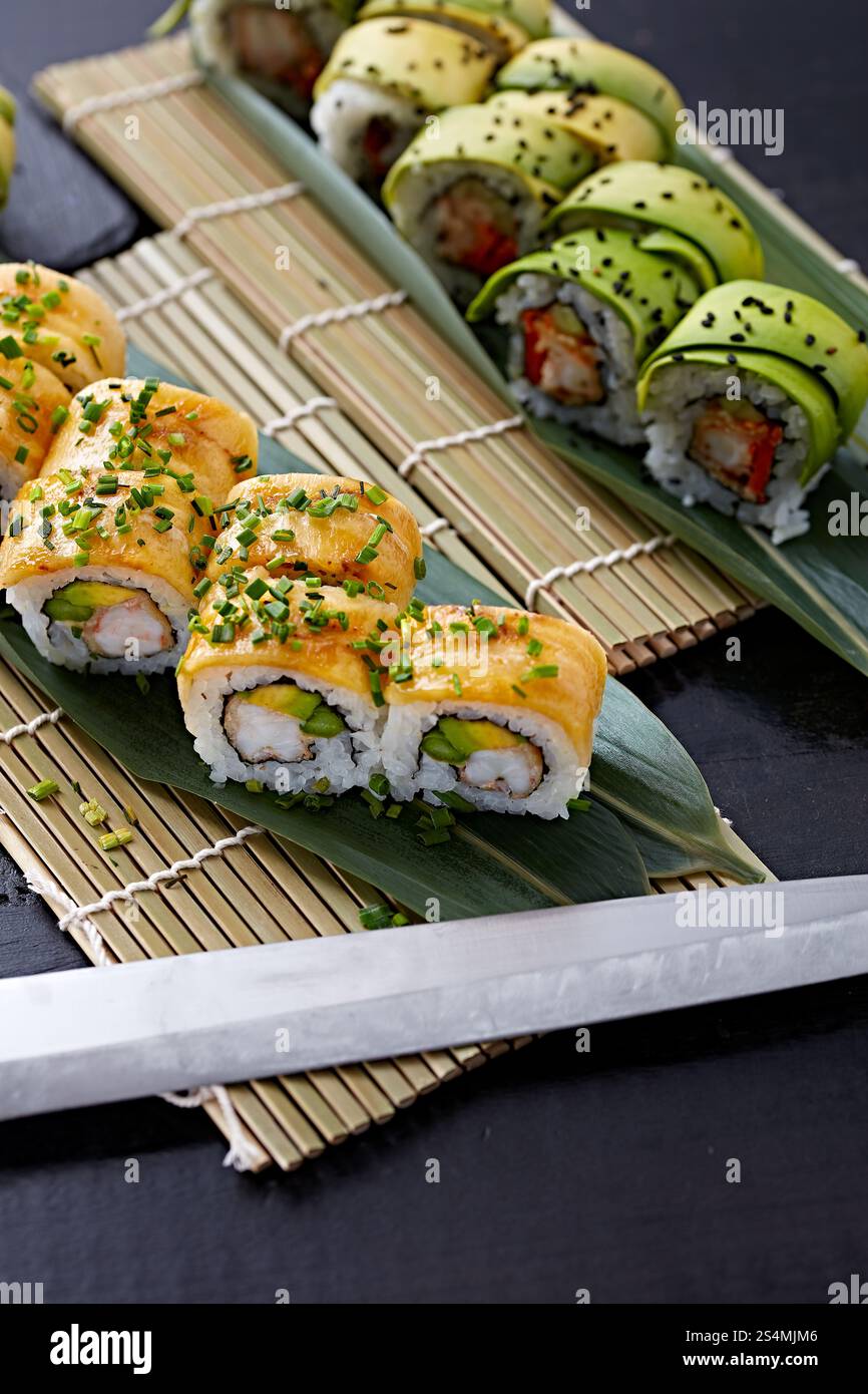 Verschiedene Sushi-Brötchen, elegant auf einer Bambusmatte platziert, mit Avocado- und Meeresfrüchtebelag, garniert mit Sesamsamen und grünen Zwiebeln. Stockfoto