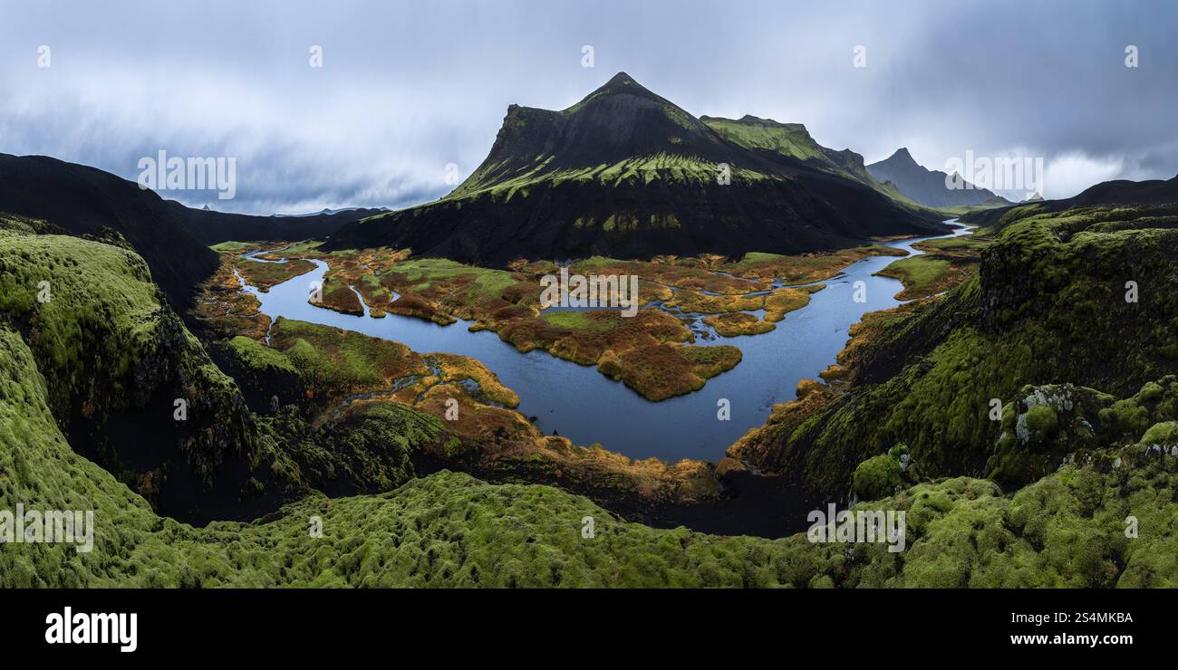 Ein atemberaubender Panoramablick auf einen moosbedeckten vulkanischen Berg mit einem sich windenden Fluss und farbenfrohen Feuchtgebieten im isländischen Hochland mit Dramat Stockfoto