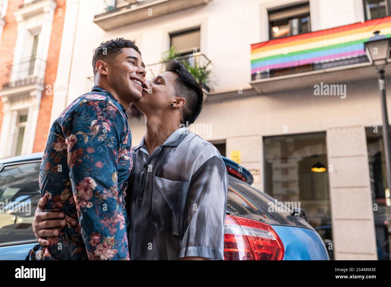 Ein fröhliches lateinisches Schwulenpaar teilt einen zärtlichen Moment auf einer lebhaften Straße in Madrid mit einer Regenbogenfahne im Hintergrund, die Stolz und Liebe symbolisiert. Stockfoto