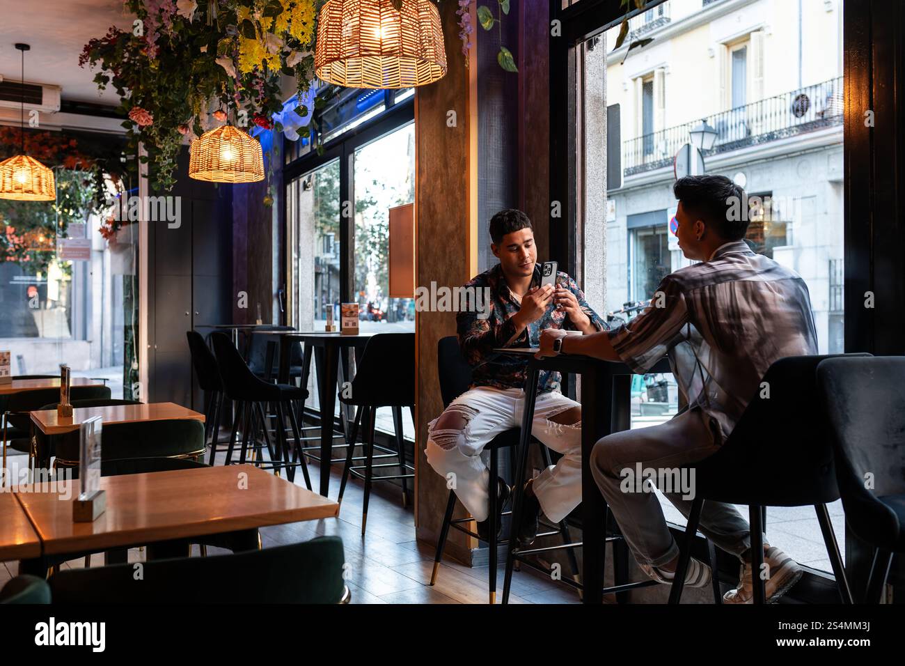 Ein lateinamerikanisches Schwulenpaar, das einen Moment in einem stilvollen Café genießt, umgeben von schickem Interieur und warmer Beleuchtung, die ein gemütliches, aber modernes urbanes Ambiente hervorhebt. Stockfoto