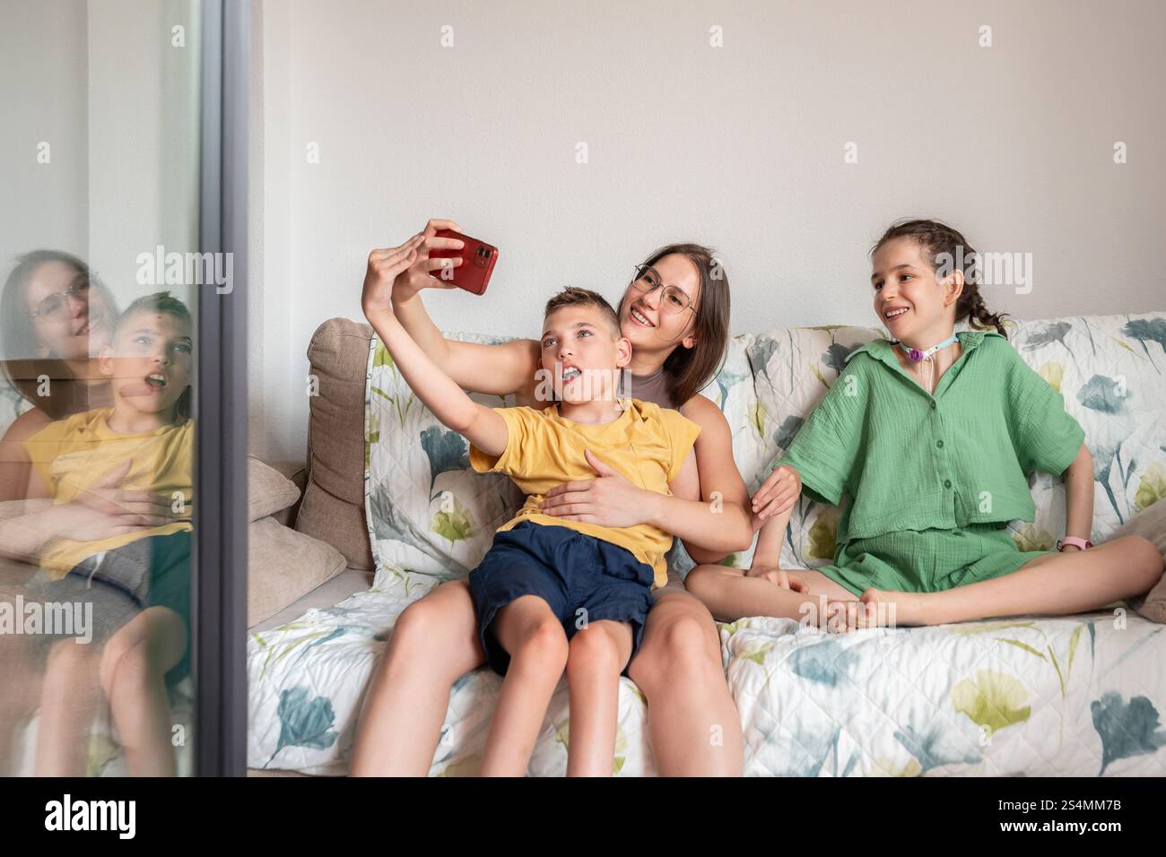 Eine fröhliche Familie teilt sich einen liebevollen Moment auf der Couch und nimmt ein Selfie auf. Zur Familie gehören ein Mädchen mit Tracheostomie und ein Junge mit Zerebralparese. Stockfoto