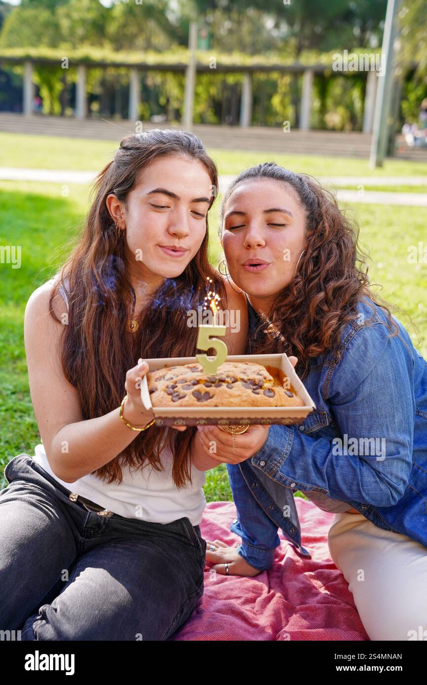 Ein romantisches junges Frauenpaar bläst eine „5“ Kerze auf einer Geburtstagskuche aus, während es auf einer Picknickdecke in einem sonnigen Park sitzt, was ein Beispiel für Freude ist Stockfoto