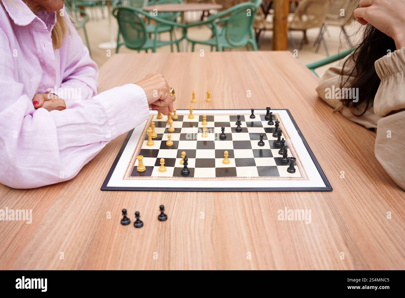 Mutter und Tochter spielen draußen auf einem Holztisch ein Schachspiel. Der Fokus liegt auf der strategischen Bewegung des Spielers in rosa grünen Stühlen und lässig Stockfoto