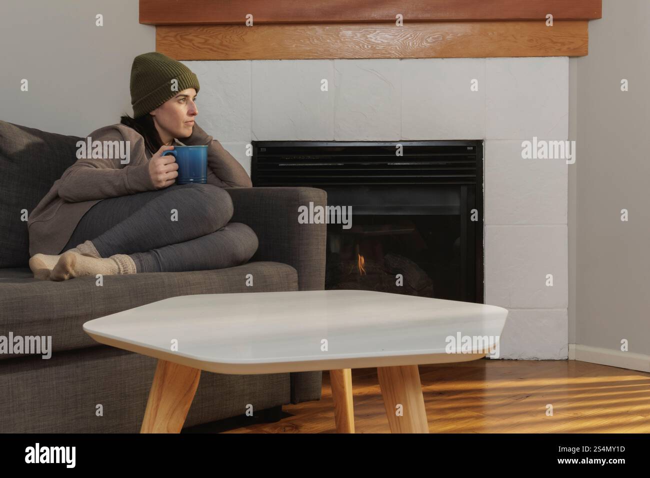 Frau saß auf einer Couch neben dem Kamin und hielt einen Becher Stockfoto