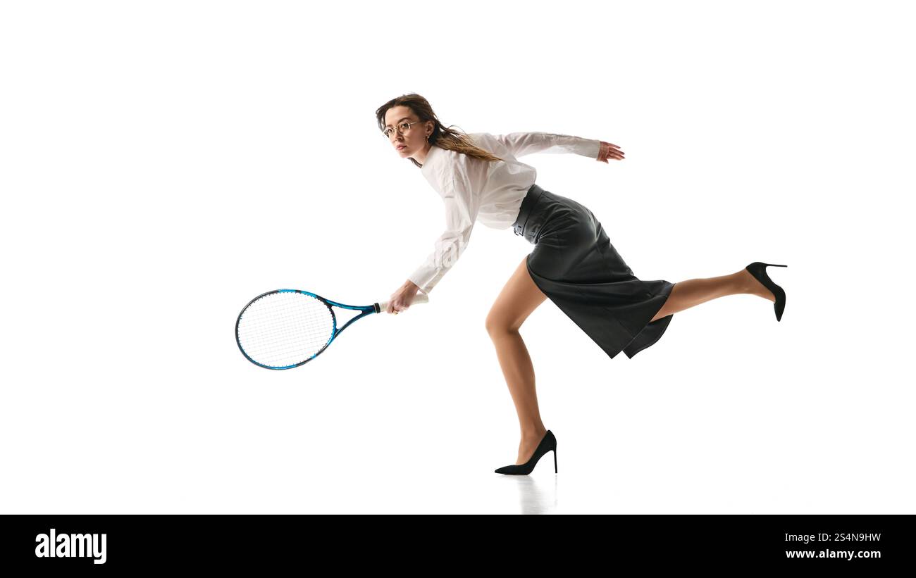 Entschlossene Frau in schwarzem Lederrock und weißer Bluse stürzt mit Tennisschläger nach vorne und verkörpert Agilität und Fokus auf weißes Studio Stockfoto