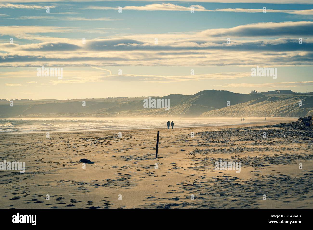 Eine entfernte Silhouette am Strand, bei der die Sonne unter dem Himmel ein goldenes Licht über Cruden Bay Beach in Aberdeenshire, Schottland, ausstrahlt Stockfoto