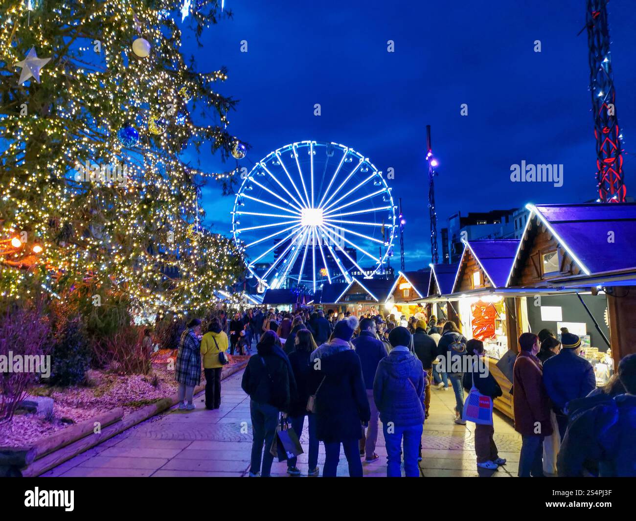 Besucher schlendern durch den lebhaften Weihnachtsmarkt am Place de Jaude, Clermont-Ferrand, der beleuchtete Verkaufsstände, ein Riesenrad und ein vib bietet - Smartphone-aufgenommenes Stockfoto