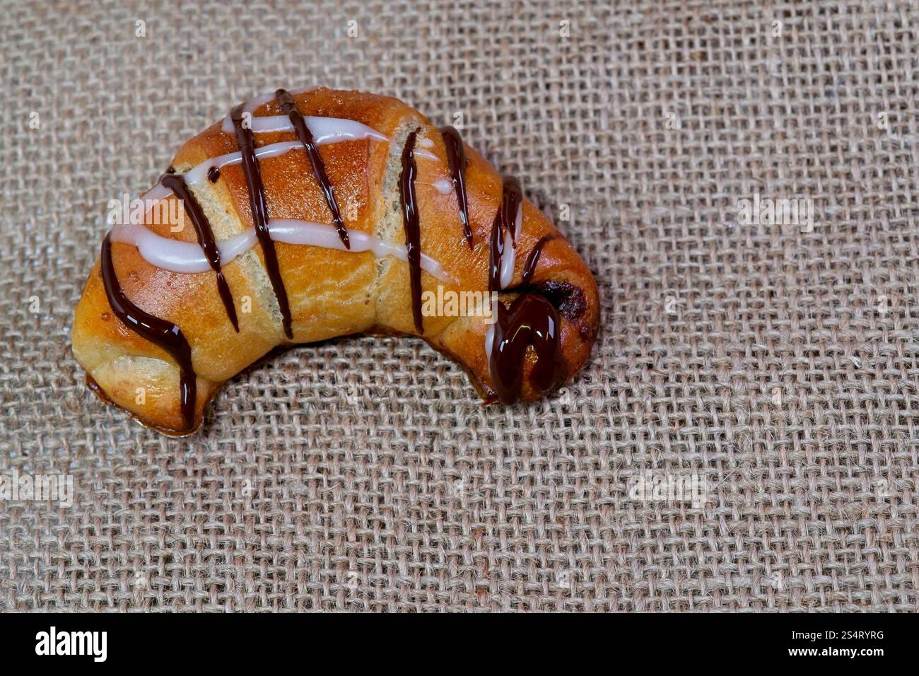 Dies ist ein Makrofoto eines einzelnen Croissants, das offen auf rustikalem Stoff präsentiert wird, der mit Schokolade und Glasur verziert ist, in einem negativen Raum. Stockfoto