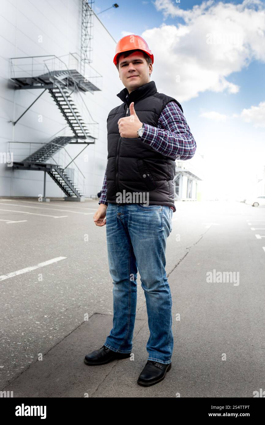 In voller Länge Portrait von lächelnden Arbeiter in Schutzhelm zeigt Daumen oben auf der Baustelle Stockfoto